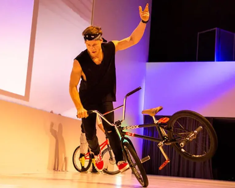 Ein Mann, der ein schwarzes Kopftuch und ein ärmelloses Hemd trägt, führt in einer Halle einen Trick auf einem BMX-Rad vor, wobei er mit einer erhobenen Hand auf dem Vorderrad balanciert und das Rad zur Seite kippt, um einen Eintrag in das Künstlerverzeichnis zu erhalten.