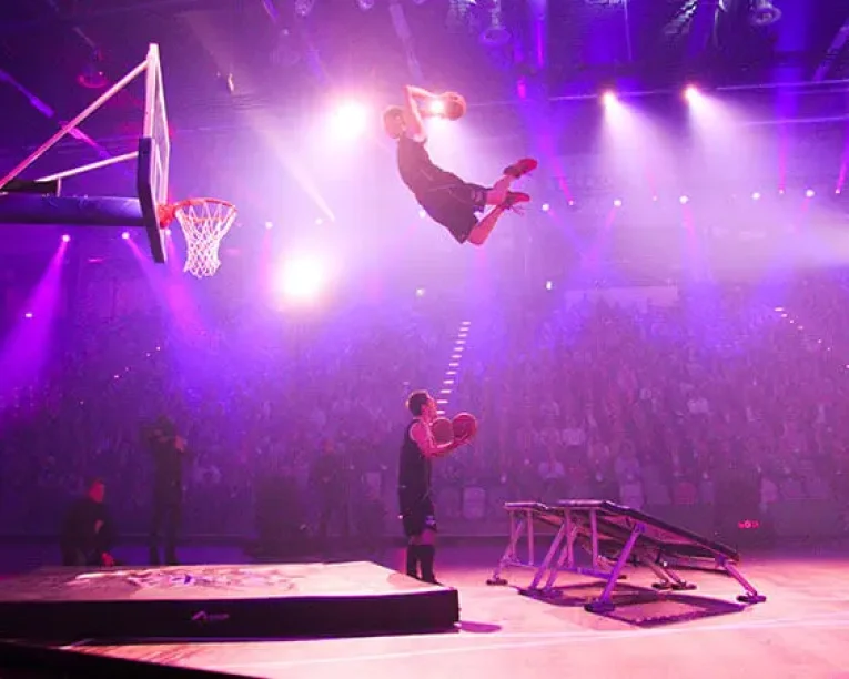 Ein Basketballspieler fliegt durch die Luft in Richtung Korb, nachdem er für eine aufregende Slam Dunk Show von einem Trampolin gesprungen ist, und hält dabei einen Ball in der Luft, während eine andere Person unter rosa und lila Bühnenlicht vor einem großen Publikum zusieht.