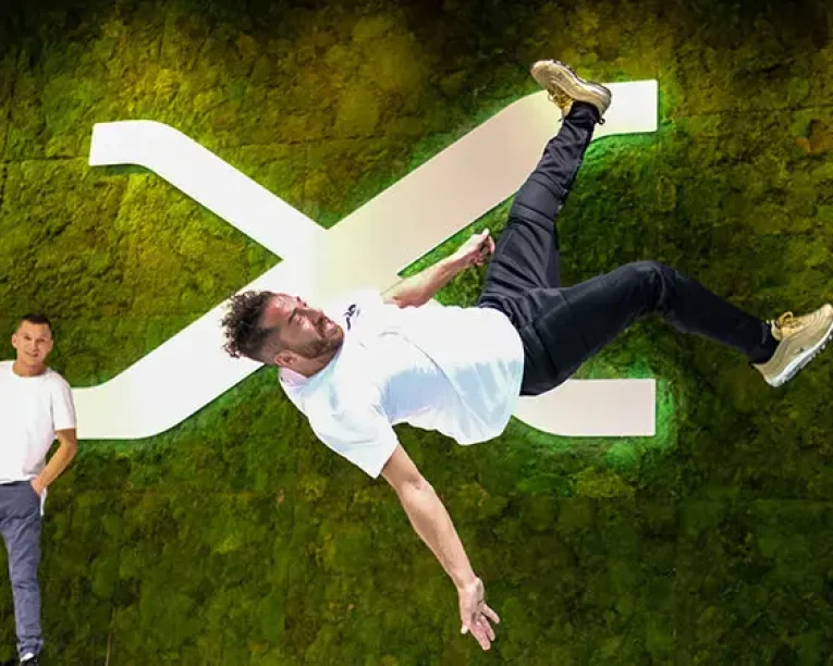 Ein Freestyle-Künstler in weißem Hemd und Jeans vollführt einen Salto in der Luft vor einer grünen Mooswand mit einem großen weißen X, während ein anderer Mann im Hintergrund zusieht.