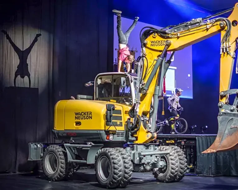 Ein Darsteller macht einen Handstand auf dem angehobenen Arm eines großen gelben Baggers auf der Bühne während einer Urban Street Show, mit dramatischer Beleuchtung und einem Schattenwurf auf einen schwarzen Vorhang. Andere Darsteller und Bühnenrequisiten sind in der Nähe zu sehen.