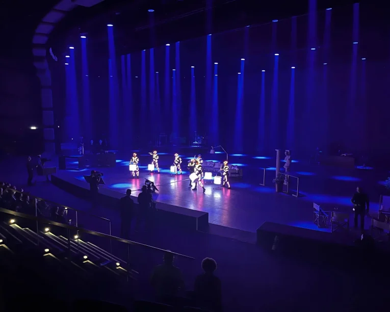 Auf einer mit blauen Scheinwerfern beleuchteten Bühne führt eine kleine Gruppe von Darstellern in weißen Kostümen eine elektrisierende LED-Trommel-Show auf, während mehrere Personen in der Nähe stehen. Das Publikum sitzt in Reihen in einem dunklen Theater.