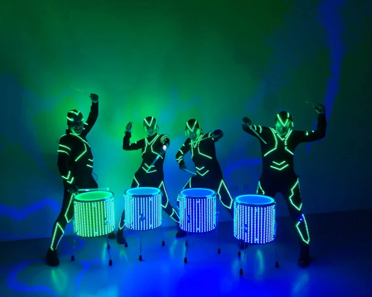 Vier Personen in futuristischen Anzügen mit leuchtenden Neonröhren führen eine dynamische LED-Trommelshow auf, bei der sie in einem dunklen Raum mit grünen und blauen Lichteffekten auf LED-beleuchteten Trommeln spielen.