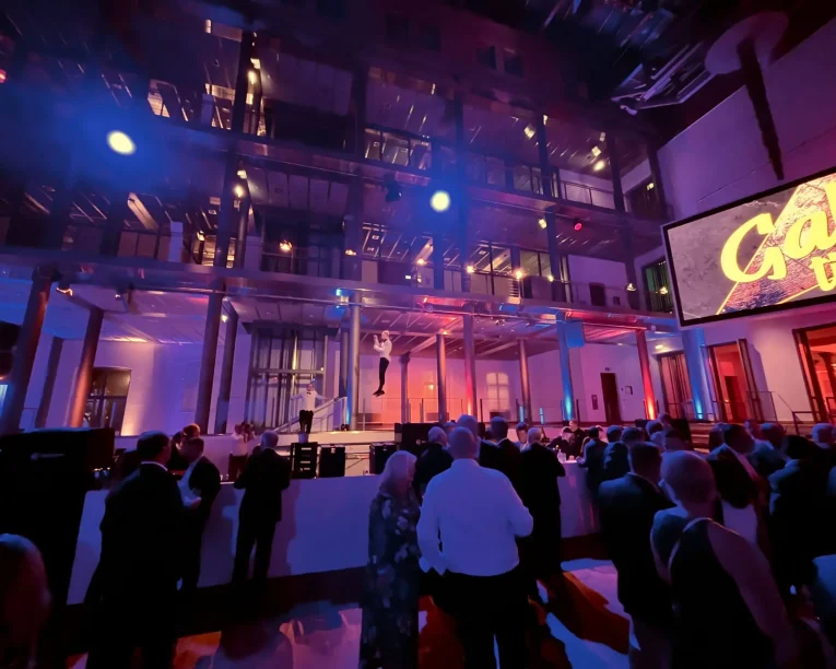 Eine große Menschenmenge in förmlicher Kleidung verfolgt den Auftritt eines Freestyle-Künstlers auf der Bühne eines modernen, hell erleuchteten Veranstaltungsortes mit bunten Lichtern. Auf einem Bildschirm rechts wird das Wort Gala angezeigt. Die Atmosphäre ist lebhaft und festlich.