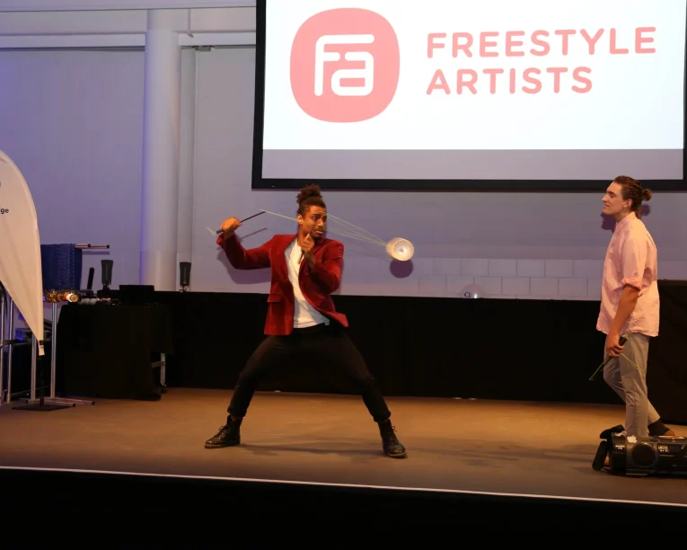 Zwei Künstler stehen auf der Bühne für eine Duo-Diabolo-Show. Der eine, in einer roten Jacke, wirbelt ein Diabolo, während der andere, in einem rosa Hemd, zuschaut. Hinter ihnen ist eine Leinwand mit Freestyle Artists und einem Logo sowie einem Volkswagen Banner zu sehen.