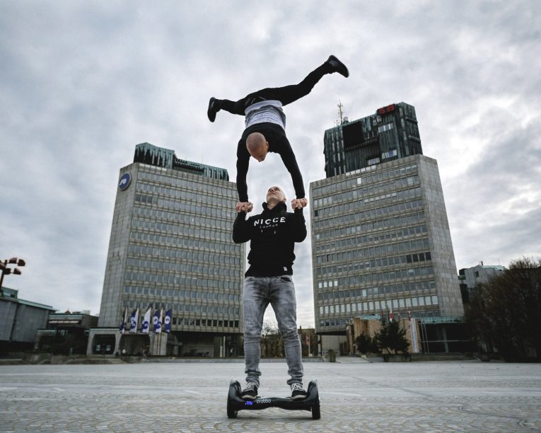 Zwei Personen vollführen im Freien einen akrobatischen Stunt, wobei eine Person auf einem Hoverboard balanciert und die andere kopfüber über ihren Kopf hebt, vor dem Hintergrund moderner Bürogebäude.