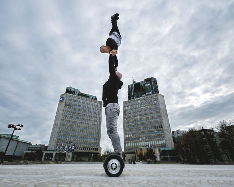 Zwei Personen vollführen auf einem städtischen Platz einen akrobatischen Balanceakt auf einem Einrad, wobei eine Person die andere kopfüber festhält. Im Hintergrund sind hohe Gebäude und ein bewölkter Himmel zu sehen.