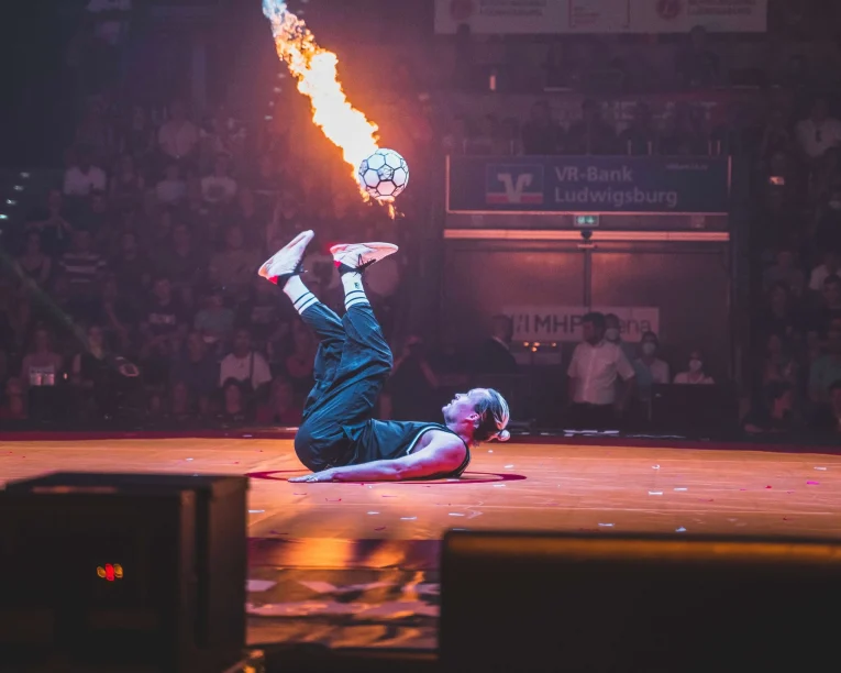 Ein Fußball-Freestyler liegt auf dem Rücken auf der Bühne und schießt einen Fußball in die Luft, aus dem Flammen schießen, während das staunende Publikum im Hintergrund zusieht.