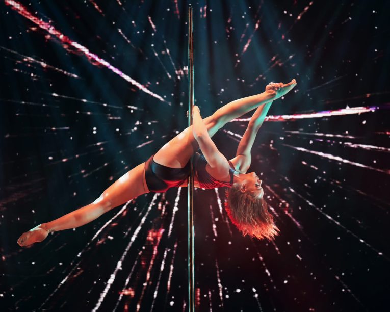 Eine Frau führt eine akrobatische Pose auf einer Stange aus, wobei sie ein Bein nach oben streckt. Sie ist umgeben von einem dramatischen Lichteffekt im Hintergrund, der eine dynamische und energiegeladene Szene erzeugt.