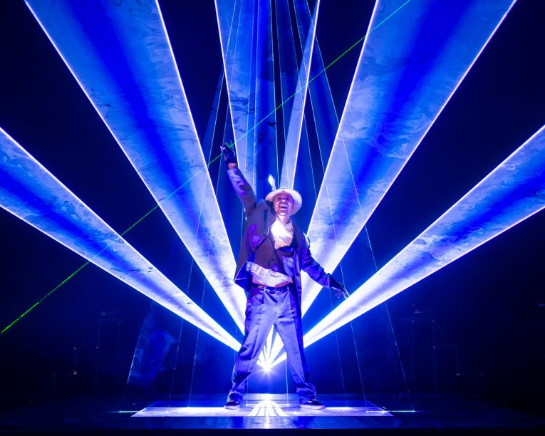 Laser Dance Show .Ein Darsteller mit Hut und Jacke steht auf der Bühne und wird von hellen blauen und weißen Laserstrahlen beleuchtet, die sich hinter ihm und um ihn herum ausbreiten und einen dramatischen und lebendigen visuellen Effekt erzeugen.