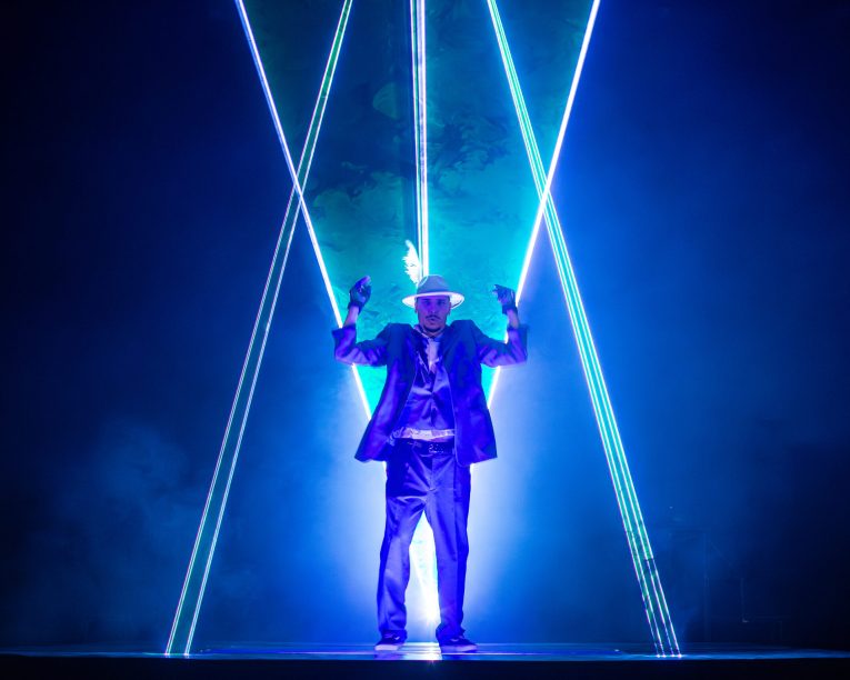 Laser Dance Show .Eine Person in Anzug und Hut steht auf der Bühne, umgeben von sich kreuzenden blauen und weißen Laserstrahlen, mit einem dramatischen blauen Leuchten und Rauch im Hintergrund.