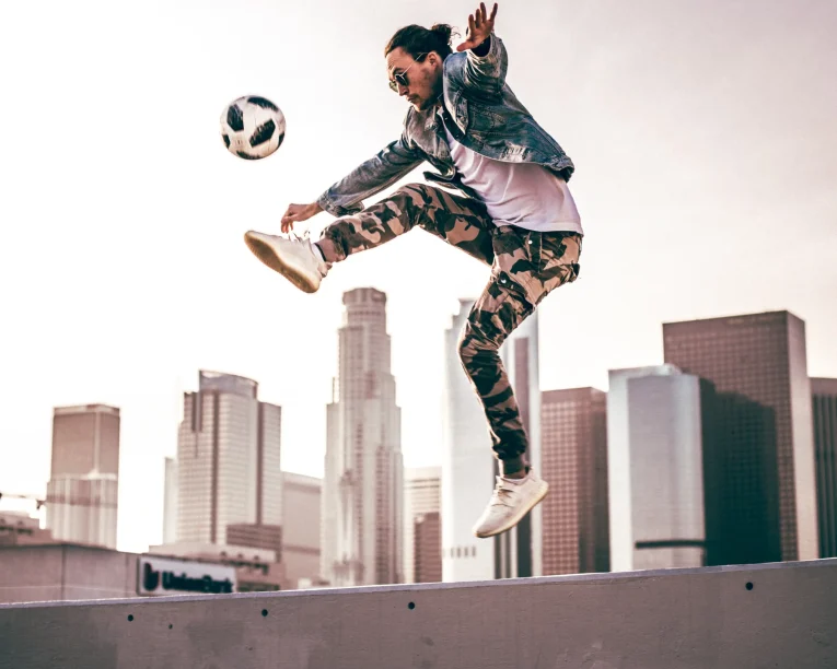 Ein Fußball-Freestyler in Jeansjacke und Camouflage-Hose springt in die Luft, während er einen Fußball kickt, mit hohen Stadtgebäuden und einem rosafarbenen Himmel im Hintergrund.