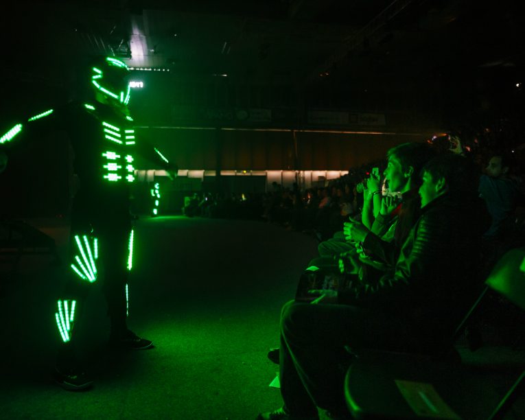 Ein Performer, der einen Anzug trägt, der mit leuchtenden grünen LED-Lichtern bedeckt ist, steht vor einem Publikum, das in einem abgedunkelten Raum sitzt, und beleuchtet den Raum mit leuchtenden Neonmustern.