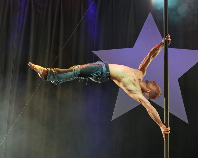 Ein hemdsärmeliger Mann in Jeans führt einen horizontalen Griff an einer vertikalen Stange auf der Bühne aus, mit einem großen Sternenhintergrund hinter ihm und dramatischer Bühnenbeleuchtung.