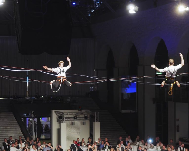 Zwei Performer in Trapezen balancieren auf parallelen Seilen hoch über einem großen, sitzenden Publikum in einer geräumigen, mit Bögen gefüllten Halle, wobei Scheinwerfer die Szene beleuchten.