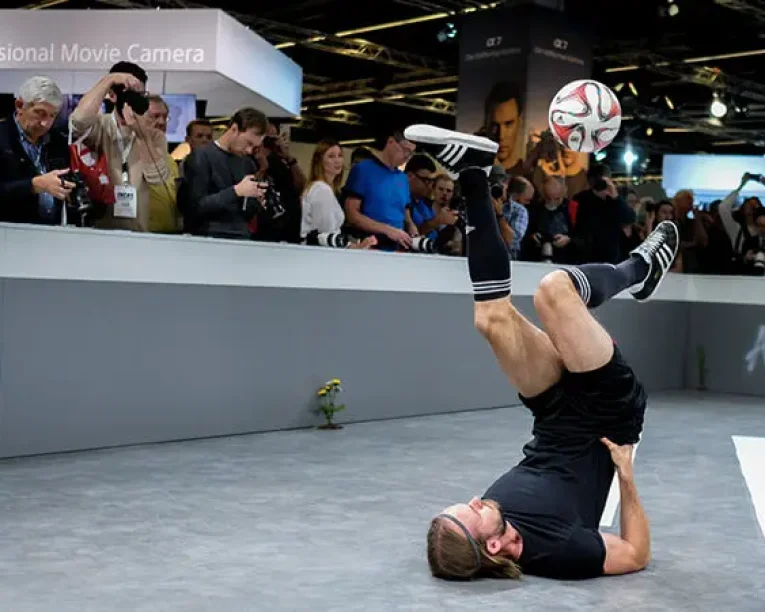 Ein Mann führt einen Freestyle-Fußballtrick auf dem Rücken vor, wobei er einen Fußball auf dem Fuß balanciert, und fesselt die Menge mit seinen beeindruckenden Messeshows, während die Leute in einer Halle zusehen und Fotos machen.