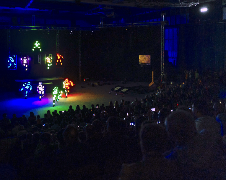 Ein großes Publikum beobachtet Darsteller in leuchtenden, bunten LED-Anzügen auf einer dunklen Bühne während einer Indoor-Veranstaltung. Die Bühne ist hell mit Neonröhren beleuchtet, während der Rest des Raums schummrig ist. Viele Zuschauer filmen.