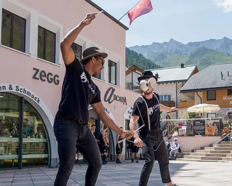 Zwei Straßenkünstler in schwarzen Hemden und Hüten unterhalten mit Jonglage, Tricks und einem Hauch von Basketball-Dunking-Show-Flair auf einem belebten Schweizer Stadtplatz, umgeben von Geschäften und Bergen unter einem sonnigen Himmel.