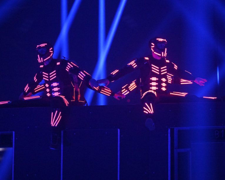 Zwei Performer in leuchtenden, futuristischen LED-Anzügen sitzen während einer Live-Show mit ausgestreckten Beinen auf einem Podest, umgeben von blauer Bühnenbeleuchtung und Strahlen.