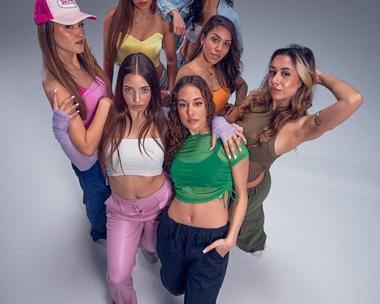 Sieben junge Frauen posieren selbstbewusst in bunten, trendigen Outfits vor einem schlichten, hellen Hintergrund. Einige stehen mit den Händen auf den Hüften oder in den Taschen, während eine eine rosa Kappe trägt. Alle blicken direkt in die Kamera.