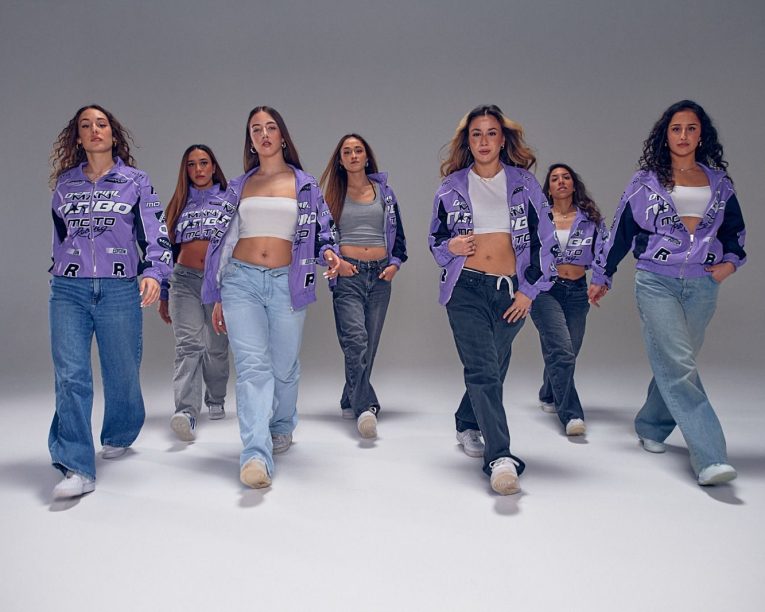 Sieben Frauen gehen in einem Studio selbstbewusst auf die Kamera zu. Sie tragen passende lilafarbene Jacken, Crop-Tops und Jeans, haben einen neutralen Gesichtsausdruck und nehmen lässige Posen ein. Der Hintergrund ist schlicht und sanft beleuchtet.