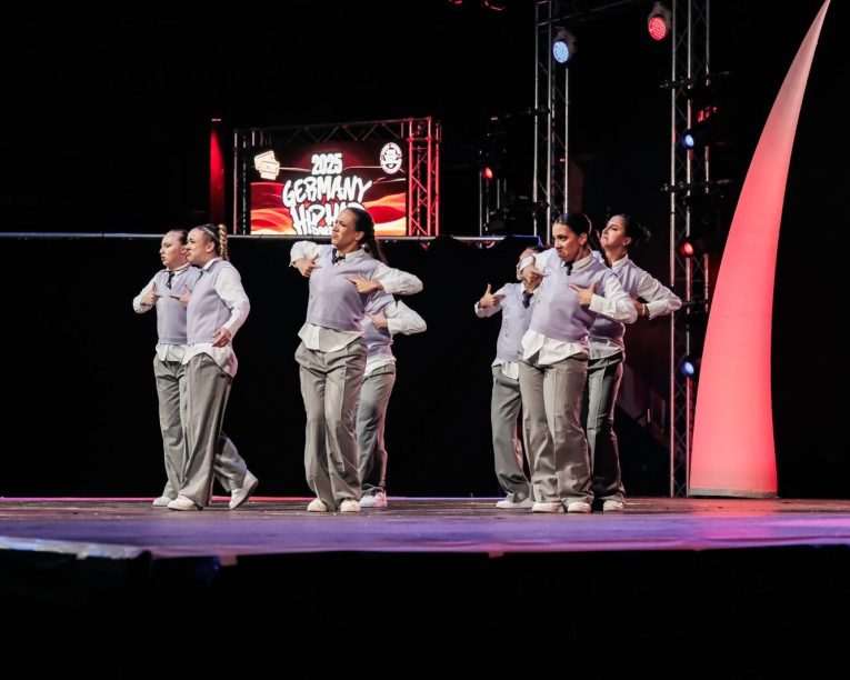 Eine Gruppe von Tänzerinnen und Tänzern in passenden grau-weißen Outfits führt auf der Bühne des Hip-Hop-Events 2023 Deutschland eine synchrone Nummer auf, mit bunten Lichtern und einem Schild im Hintergrund.