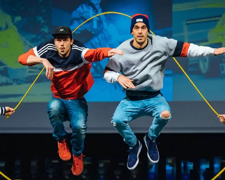 Vier Männer in legerer Streetwear springen bei einer schwungvollen Ropeskipping-Show im Gleichschritt mit einem gelben Springseil auf der Bühne, während im Hintergrund Sportbilder auf die Leinwand projiziert werden.