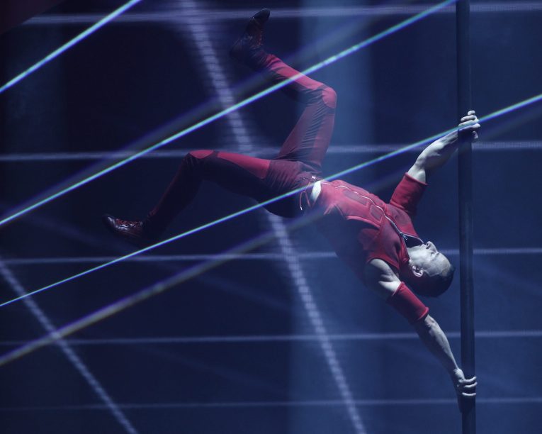 Ein Darsteller in rotem Outfit hält eine vertikale Stange mit ausgestreckten Beinen horizontal in der Luft, umgeben von dramatischer Bühnenbeleuchtung und Laserstrahlen.