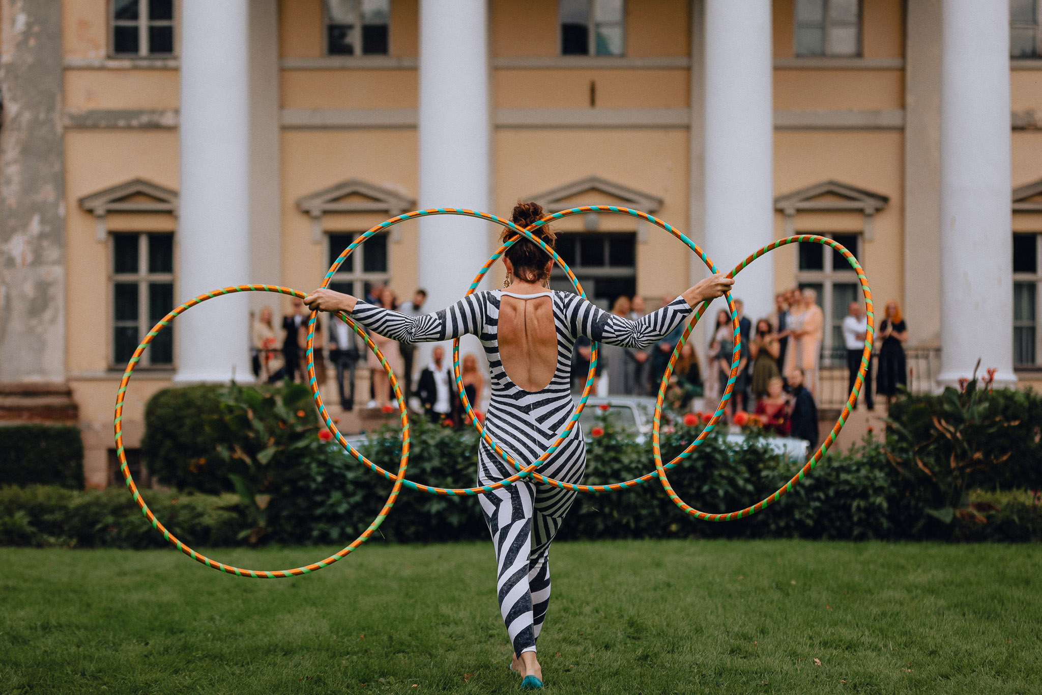 Eine Person in einem zebragestreiften Outfit steht mit dem Gesicht zur Wand im Gras und wirbelt gekonnt vier bunte Hula-Hoop-Reifen vor einem großen Gebäude mit weißen Säulen und Menschen im Hintergrund.