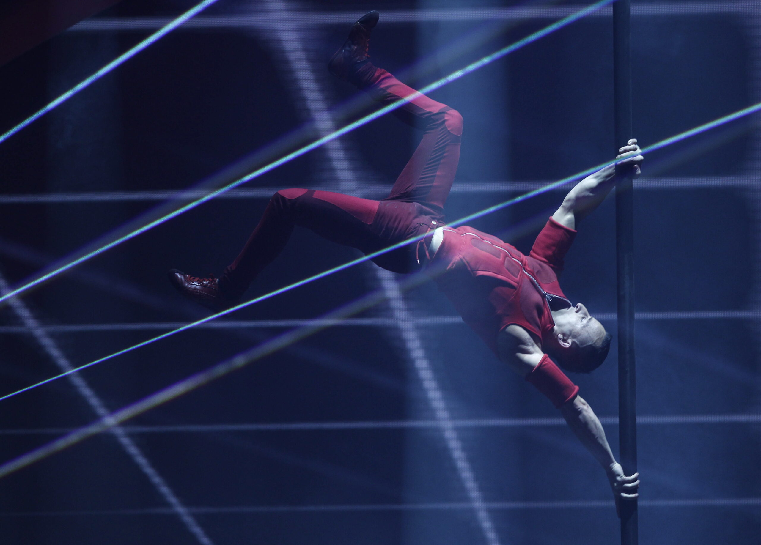 Ein Darsteller in rotem Outfit hält eine vertikale Stange mit ausgestreckten Beinen horizontal in der Luft, umgeben von dramatischer Bühnenbeleuchtung und Laserstrahlen.