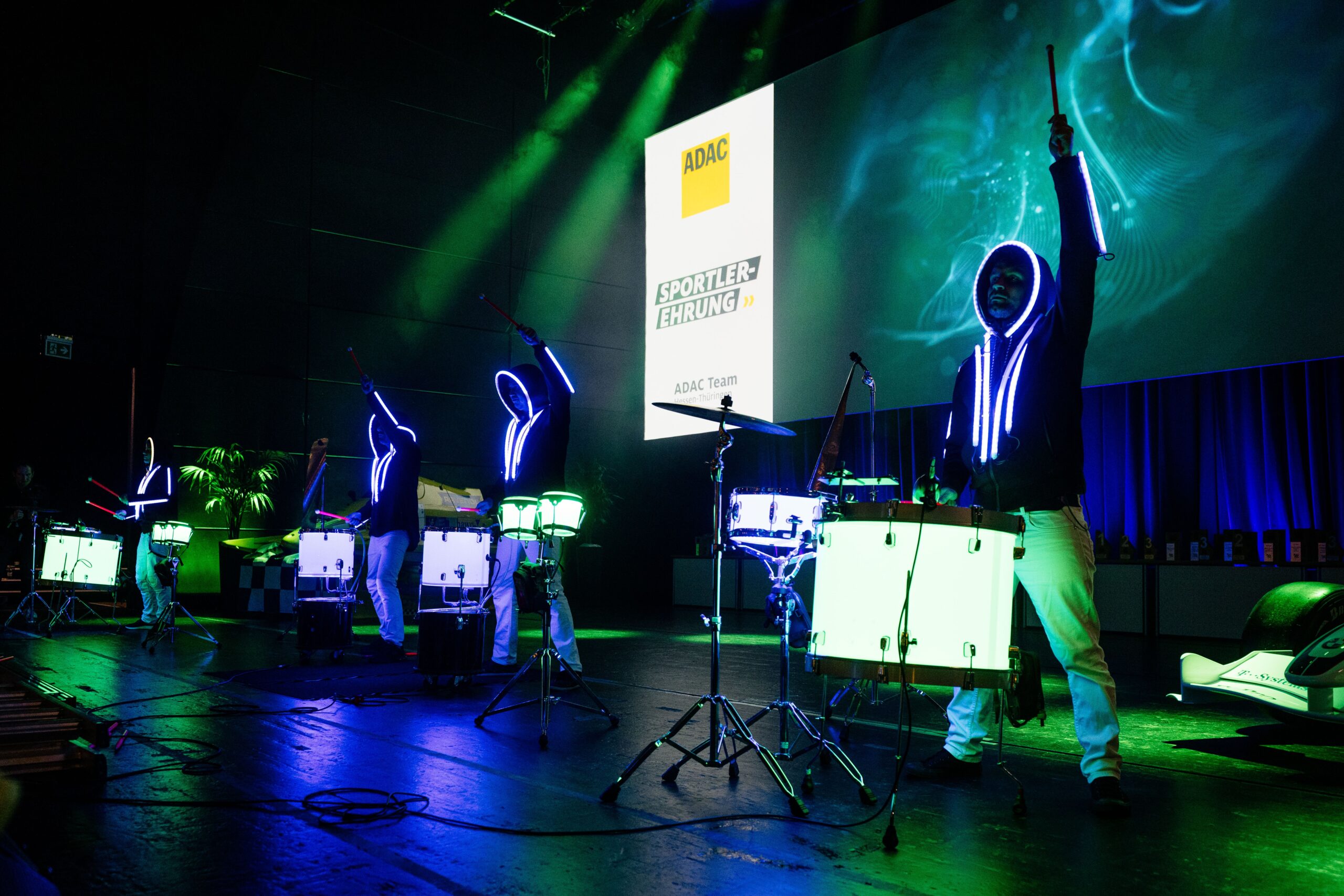 Vier Schlagzeuger in leuchtenden Anzügen treten bei einer Indoor-Veranstaltung auf der von Neonröhren beleuchteten Bühne vor einer großen Leinwand mit dem ADAC-Logo und der Sportlerehrung energisch auf.