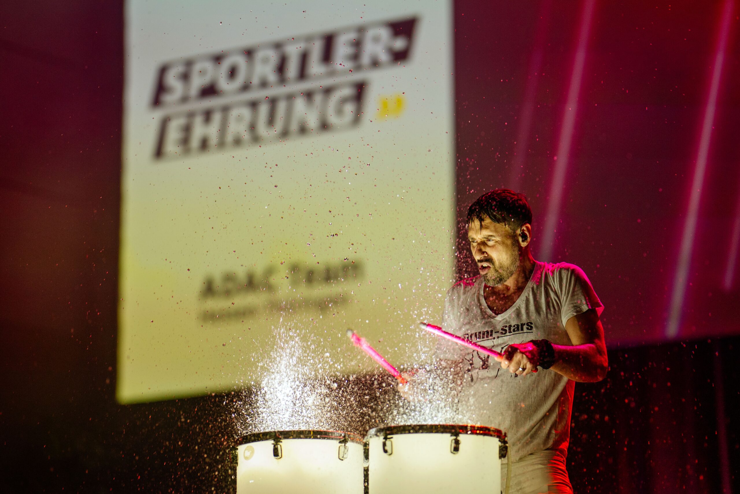 Ein Mann spielt auf der Bühne energisch auf beleuchteten Trommeln, wobei Wassertropfen durch die Luft fliegen. Auf einer großen Leinwand hinter ihm steht in fetten Lettern SPORTLER-EHRUNG und ADAC Team.